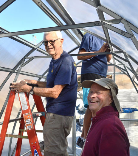 Two men smiling and working together to assemble a greenhouse frame.